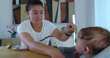 Anne bebeğini kaşıkla dikkatlice besliyor, yüksek bir sandalyeye oturmuş, evde yemek yerken sessiz bir bağ anının tadını çıkarıyor.