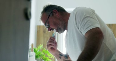 Adam lavaboda dişlerini fırçalıyor, hafifçe öne eğiliyor, temiz ve modern banyoda sabah rutininin bir parçası olarak kendine bakım ve ağız temizliğine odaklanıyor.