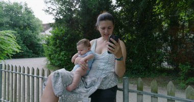 Anne bir video görüşmesine odaklanırken bebeği kucağına alıyor, ikisi de açık havada yemyeşil bir yerde oturuyor, anne akıllı telefonuyla konuşurken bebek sakince dinleniyor.