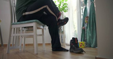 Babam bir sandalyede oturmuş botlarını bağlarken bir bebek elinde renkli bir oyuncak bloğuyla perdelerin yanında duruyor. Ahşap zemin ve doğal ışık olan bir aile yemek odasında.