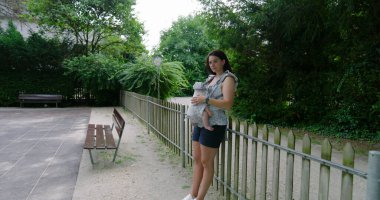 Anne, bebeği bir taşıyıcıda bebeğiyle parkta sessizce duruyor. Yeşilliğin ortasında huzurlu bir yansımanın tadını çıkarıyor. Doğayla olan bağını ve sakinliğini simgeliyor.