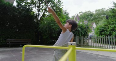 Çocuk sarı bir atlıkarıncada dikilirken elinde sopayla arka planda yeşillik ve çitle