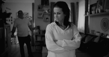 Tartışma sırasında kollarını çapraz uzatmış bir kadın, arka planda yürüyen ve el kol hareketi yapan bir adam, gergin ve duygusal ilişki çatışması, siyah ve beyaz