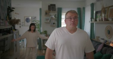 Erkek, arka plandaki kadınla tartışırken gözle görülür şekilde üzgün, oturma odasında ilişki gerilimi, duygusal ve dramatik anlaşmazlık anı