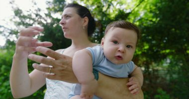 Yeşil bir parkta annesi tarafından tutulan bebek, şaşkın ve meraklı bir ifadeyle, doğal bir çevrede keşif ve keşif anını yakalıyor.