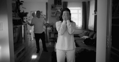 Kadın hararetli tartışmalar sırasında kafasını tutarken, arka plandaki erkek öfkeyle işaret ediyor, yoğun çatışma ve duygusal sıkıntı çekiyor..