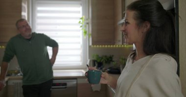 İki yetişkin modern bir mutfakta neşeli bir şekilde sohbet ediyorlar. Mavi bir kupa tutan ve gülümseyen bir kadın. Tezgaha yaslanan bir adam, rahat bir atmosfer.