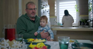 Babam yemek masasında renkli yemek ve içeceklerle bebeği tutarken annem de mutfakta, sıcak aile ortamında sıcak aile ortamını hazırlıyor.