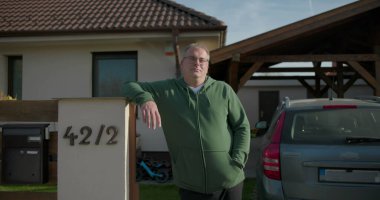 Banliyöde evi olan bir posta kutusunun yanında çitlere yaslanan ve arka planda park edilmiş bir araba olan bir adam, sakin ve ulaşılabilir bir varlık sergiliyor.