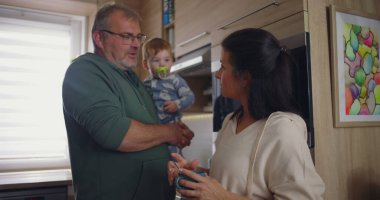 Anne modern mutfakta kahvesini karıştırırken baba kucağında. Ev ortamında sakin bir aile ortamının altını çiziyor.