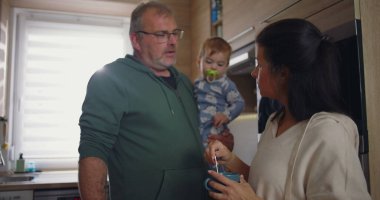 Anne modern mutfakta kahvesini karıştırırken baba kucağında. Ev ortamında sakin bir aile ortamının altını çiziyor.