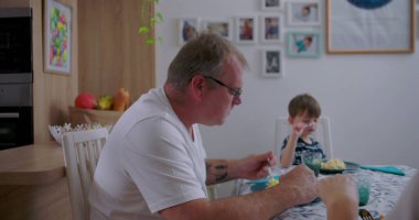 Baba ve küçük oğlu mutfakta öğle yemeği yiyorlar, sıradan aile anları, yemek servisi yapan babaya odaklan, çocuk dikkatle dinliyor, rahat bir atmosfer.