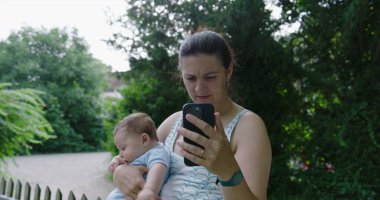 Anne akıllı telefonuyla bir video görüşmesi yaparken bebeği kucağında tutuyor, anne yaz günü sohbete odaklanıyor.