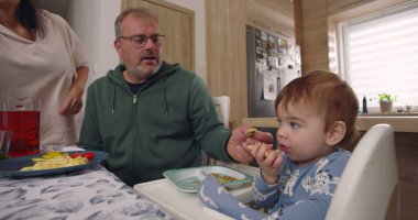 Baba yemek sırasında bebek maşasında oturur, anne tabağın yanında durur, aile bağlarını yemek masasında kurar, çocuk yemek yer.