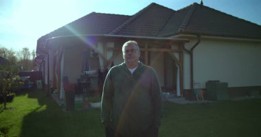 Evin arka bahçesinde duran adam, sakin bir ifade, banliyö manzarası, arka planda yeşil çimenler ve veranda, sakin atmosfer, rahat ve rahat bir an.