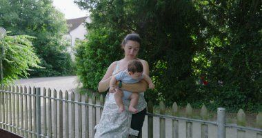 Anne dışarıda bebeğini tutuyor, çocuğuyla meşgul olurken gülümsüyor, arka planda yeşillikler ve ağaçlar var, doğal park ortamında neşeli ve hassas bir an.