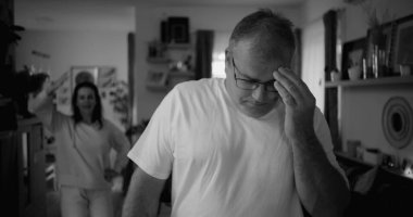 Tartışma sırasında yoğun ifadeli erkek portresi, oturma odasında arka planda kadın, duygusal gerilim ve gergin ilişki dinamiklerine odaklan, siyah-beyaz