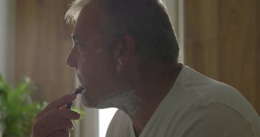 Jiletle tıraş olan, başını hafifçe eğen, temiz bir tıraş için baskı uygulayan, yüzünde köpük olan, arka planda banyo detayları, hijyen ve bakım yapan bir adam.