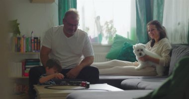 Baba ve çocuk sehpanın üzerinde oyuncak trenle etkileşime giriyor, anne arka planda beyaz köpeği tutuyor, yakın aile bağı ve kapalı alanda kaynaşma.