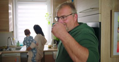 Adam elinde fincanla gülümserken, anne mutfakta bebeği tutuyor, neşeli aile anları günlük ev rutinlerinde, gündelik ve sıcak etkileşimler