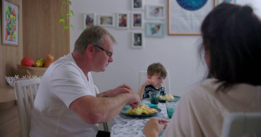 Baba ve küçük oğlu mutfakta öğle yemeği yiyorlar, sıradan aile anları, yemek servisi yapan babaya odaklan, çocuk dikkatle dinliyor, rahat bir atmosfer.