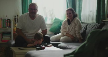 Anne ve baba kanepede oturup çocuğa gülümsüyor kahve masasında oyuncak trenle oynuyor rahat oturma odası düzeninde, neşeli aile anları, kapalı alanda kaynaşma zamanı.