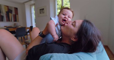 Anne sıcak ve sevgi dolu bir anda bebeği öpüyor ve kucaklıyor, doğal ışık sıcak bir oturma odasına akıyor, anne ve çocuk arasında neşeli ve samimi bir bağ kuruyor.