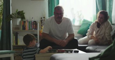 Babam, kahve masasında oyuncak trenle oynayan bir çocukla oturuyor. Anne, kanepeden gülümsüyor ve gözlemliyor. Sıcak aile bağları ve huzurlu ev ortamında samimi bir bağ.