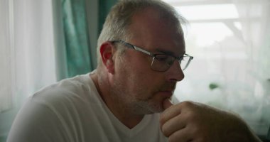 Adamın biri, çenesini elinde dinlendirirken, sakin ve rahat bir evde yumuşak yeşil perdeleri olan bir pencerenin yanında otururken düşünceli bir şekilde bakıyor.