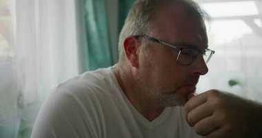 Adamın biri, çenesini elinde dinlendirirken, sakin ve rahat bir evde yumuşak yeşil perdeleri olan bir pencerenin yanında otururken düşünceli bir şekilde bakıyor.