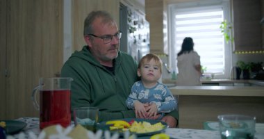 Babam yemek masasında renkli yemek ve içeceklerle bebeği tutarken annem de mutfakta, sıcak aile ortamında sıcak aile ortamını hazırlıyor.