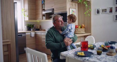 Baba kucağında bebek ve gülümsüyor. Sıcak bir mutfak ortamında neşeli ve sevecen bir an yaşıyor.