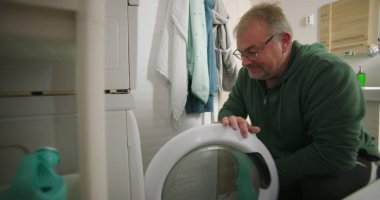 Çamaşır makinesine çamaşır sepeti ve temiz ve modern banyo ortamında havluları koymaya hazırlanırken gülümseyen bir adam.