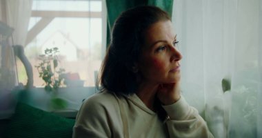 Sakin ve düşünceli bir ifadeyle dışarı bakan bir kadının yakın çekimi, çenesini elinde dinlendirirken, yumuşak perdelerle çerçevelenmiş ve rahat bir iç mekan.