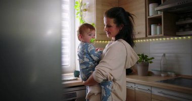 Annenin bebeği tezgahın yanında tutması, sıradan bir sabah rutini sırasında sıcak bir aile anı, evde ebeveynler ve çocuklar arasında bağ kurulması..