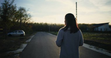 Gün batımında sakin bir yolda yürüyen bir kadın, arkasını dönmüş, huzurlu bir kırsal atmosfer, kenara park edilmiş bir araba, sakin bir dış mekan, kırsal bölgeler ele geçirilmiş