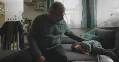 Babam, bir bebeğin yanında, emzik ile bir kanepede dinleniyor. Huzurlu bir aile salonunda, içi doğal ışıkla dolu bir el uzatıyordu.