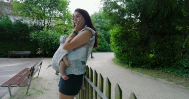 Bir taşıyıcıda bebeğini taşıyan anne, parkta dikilirken elini yüzünü dinlendirirken, bir anlık derin düşüncelere dalmış ve çevresiyle olan bağlantısını yansıtıyor.