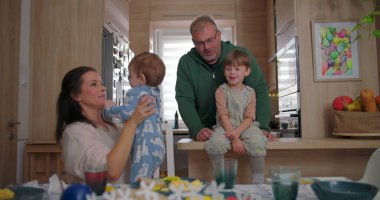 Büyük çocuk mutfak tezgahında otururken heyecanlı bir şekilde bir şeyi işaret ediyor, baba ve anne kucağında bebek, sıcak mutfakta neşeli bir aile etkileşimi.