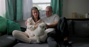 Küçük beyaz köpekle kanepede dinlenen, sakin ve samimi aile anlarının tadını çıkaran, doğal ışık ve yumuşak mobilyaları olan rahat oturma odası, sıcak atmosferi olan çift.