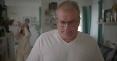 Oturma odasında hararetli tartışmalar yaşayan bir çift, görünürde üzgün duran bir adam, arka planda duygusal hareketler yapan bir kadın, ilişki çatışması sırasında gergin bir an.