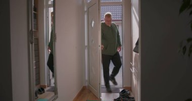 Bir adam eve ön kapıdan giriyor, küçük beyaz bir köpek tarafından karşılanıyor, ayakkabılar ve ayna yansıması ile girişte duruyor, sıcak ve sıcak bir ev ortamında.