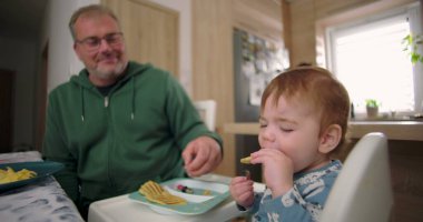 Bebek kahvaltıda yüksek sandalyede otururken waffle yiyor, baba sıcak bir ilişki kuruyor ve sıcak bir ev mutfağı kuruyor.
