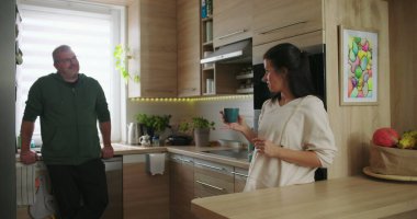 Kadın ve erkek, modern bir mutfakta rahat bir şekilde sohbet ediyorlar. Elinde kahve fincanı tutan kadınla birlikte. Parlak ve davetkar bir ortamda.