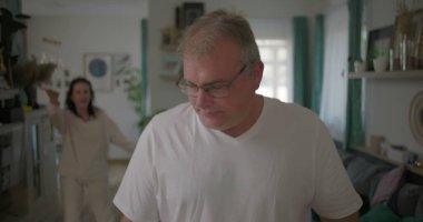 Oturma odasında hararetli tartışmalar yaşayan bir çift, görünürde üzgün duran bir adam, arka planda duygusal hareketler yapan bir kadın, ilişki çatışması sırasında gergin bir an.