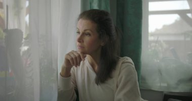 Pencerenin yanındaki rahat bir koltukta oturan kadın, kolunu yastığa koyuyor, sakin bir oturma odasında düşünceli bir ifadeyle dışarı bakıyor.