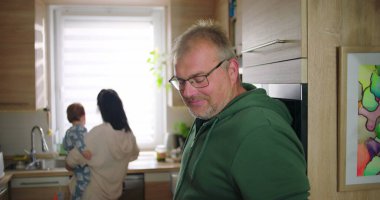 Adam elinde fincanla gülümserken, anne mutfakta bebeği tutuyor, neşeli aile anları günlük ev rutinlerinde, gündelik ve sıcak etkileşimler