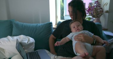 Anne kucağında bebekle evde dizüstü bilgisayarla çalışıyor. Kanepede birlikte sakin bir anın tadını çıkarıyor. Oturma odasını doğal ışık dolduruyor.