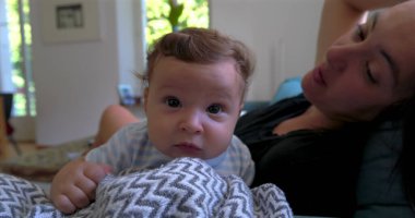 Bebek battaniyeye uzanmış merakla bakarken anne arka planda doğal ışık ve sıcak aile atmosferiyle dolu rahat bir oturma odasında gülümsüyor..