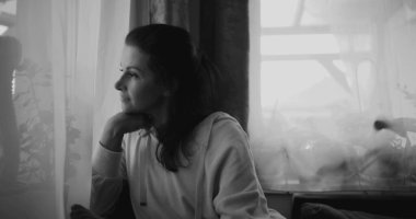 Pencereden dışarı bakan kadın, düşünceli bir duruşla çenesini dinlendiriyor, bitkilerle ve perdelerle çevrili huzurlu bir evde oturuyor, monokromatik tonları yansıtıyor.